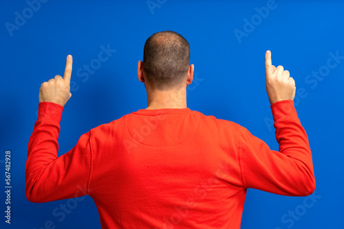 Portrait of a man with incipient alopecia from behind, raising both hands and pointing his index fingers up, isolated on a blue background.