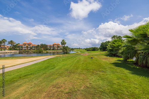 Gorgeous view of hotel building and blue water surface of pond and green grass golf field on background blue sky with white clouds. Aruba.