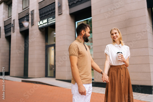 Cheerful young couple in love standing holding hands on city street and looking on each. Handsome bearded man with tattoo and cute blonde woman walking drinking takeaway coffee enjoying time together.
