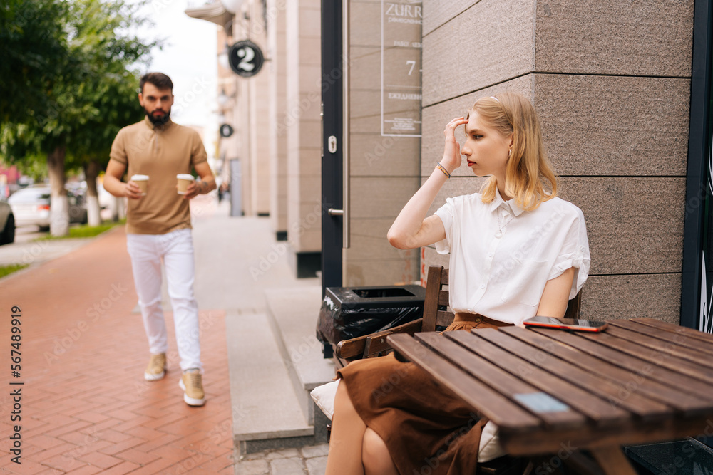 Portrait of caring young man bringing coffee to charming blonde girlfriend sitting at table in street cafe on summer day. Romantic bearded male brings hot fresh coffee to pretty woman outdoors. Portrait of caring young man bringing coffee to charming blonde girlfriend sitting at table in street cafe on summer day. Romantic bearded male brings hot fresh coffee to pretty woman outdoors.