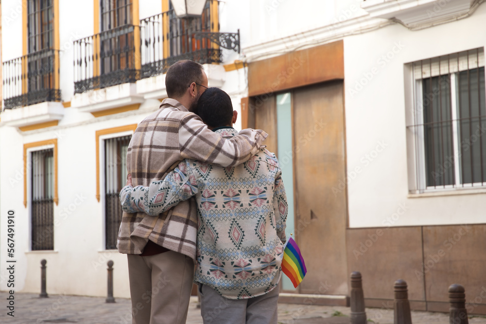 Real marriage of gay couple, hugging on the street, with a gay pride flag in their pocket and seen from the back. Concept lgtb, lgtbiq+, couples, in love, pride, hugging.