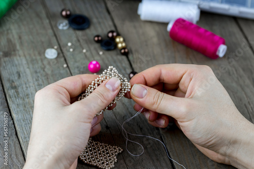 Female hands making of handmade jewellery. Box with beads, spool of threads, needle, scissors on wooden background