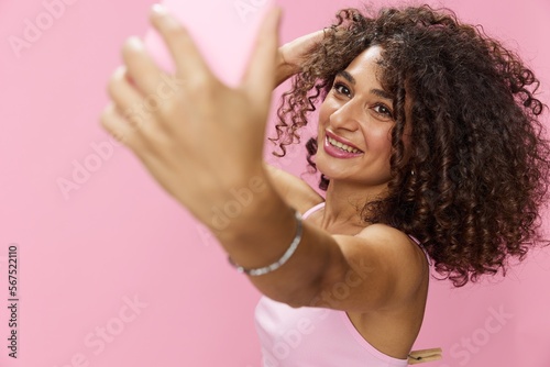 Woman blogger holding phone video call takes selfies, with curly hair in pink smile t-shirt and jeans poses on pink background, copy space, technology and social media, online
