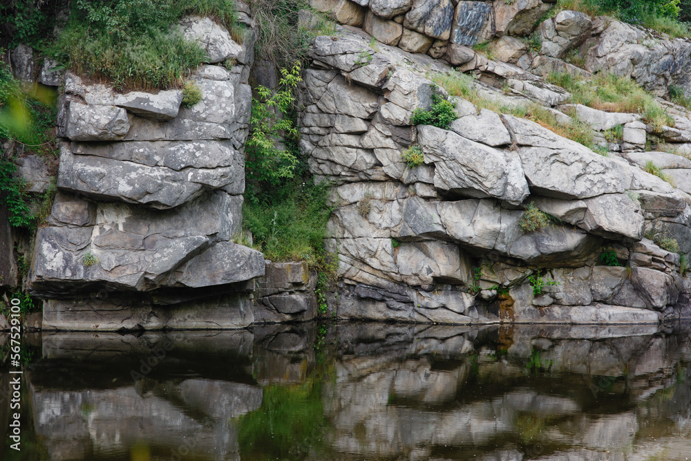 Amazing landscape with textured wall of rocks and greenery in ancient granite canyon Buky in Ukraine in summer, smooth river at the bottom, scenic view from water with colorful reflections