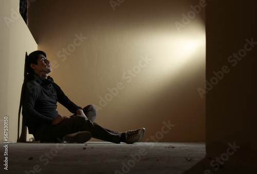 Hombre joven triste, sentado en el suelo mirando arriban y pensando con iluminación puntual hacia él , concepto de tristeza y melancolía, depresión, soledad. photo