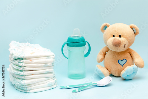 Stack of diapers with cute teddy bear toy, bottle of milk on table. set for infant newborn boy girl for baby shower present gift on blue background. Healthcare medical, hygiene concept