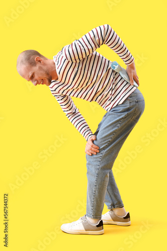 Young man warming his lower back with hot water bottle on yellow background