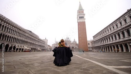Fair haired woman walks along famous empty St Marks Square holding long black dress plume and enjoying life backside view photo