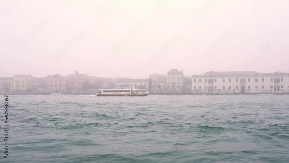 Tourist pleasure boat speeds on wide waving blue water against Venice ancient city with small buildings standing in dense fog Tourist pleasure boat speeds on wide waving blue water against Venice ancient city with small buildings standing in dense fog