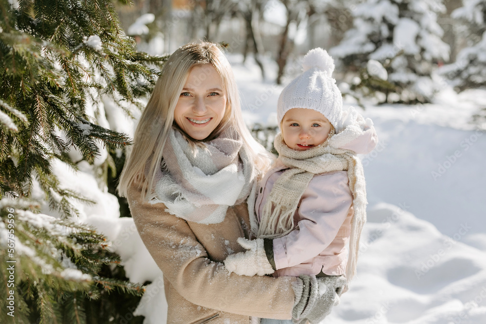 Fototapeta premium Portrait of young mom and little daughter enjoying sunny day at snowy park. Mother holding child on hands, and looking together at camera while standing in background of snowy trees