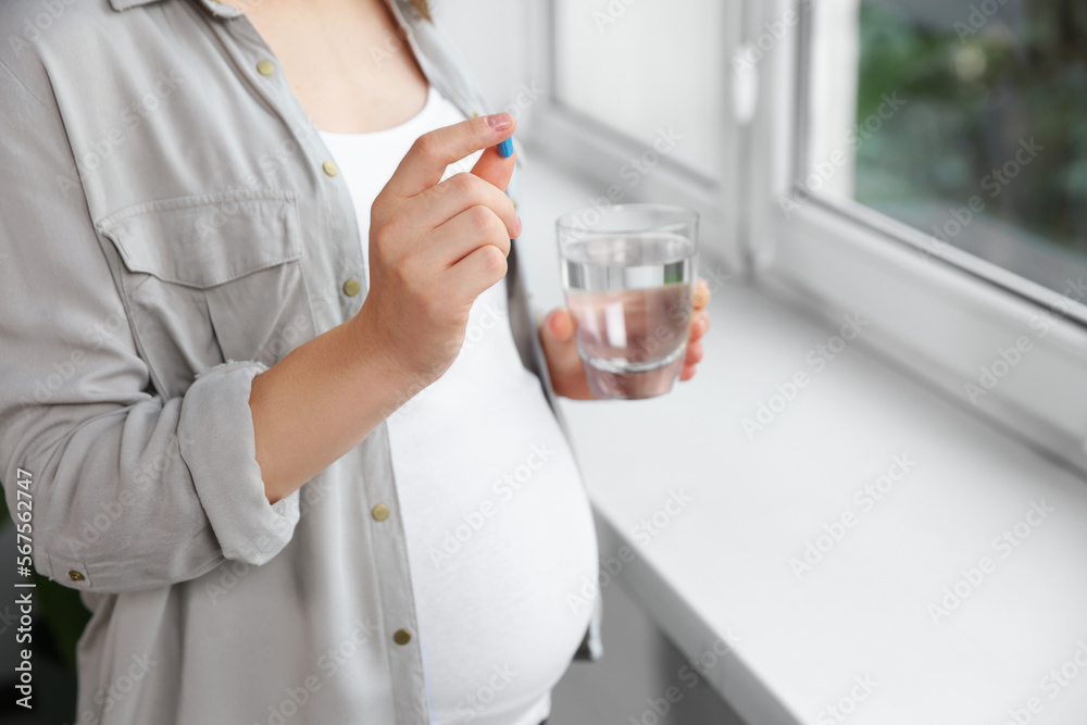 Pregnant woman holding pill and glass with water near window indoors, closeup Pregnant woman holding pill and glass with water near window indoors, closeup