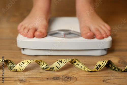 Overweight girl using scales near measuring tape on wooden floor, selective focus