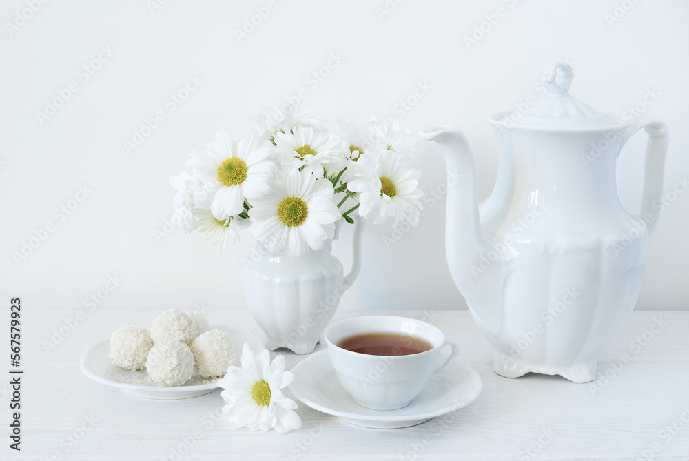 Tea and flowers. A bouquet of white flowers of daisies or chamomile in a vase on the table, a cup of tea, sweets and a teapot on a white background Tea and flowers. A bouquet of white flowers of daisies or chamomile in a vase on the table, a cup of tea, sweets and a teapot on a white background