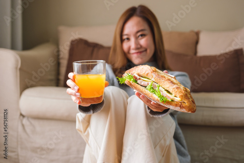 Portrait image of a young woman holding and eating french baguette sandwich and orange juice at home