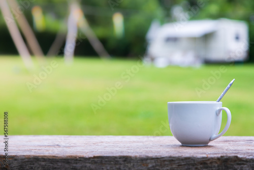 A morning cup of coffee on a wooden table overlooking the blurry tents. with bokeh blur from the trees and green lawn in the background.