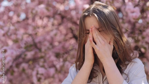 Irritated caucasian young woman standing near blooming sakura tree and scratching itchy eyes during spring time outdoors. Allergic reaction to pollen. For Advertisement of a medicinal product