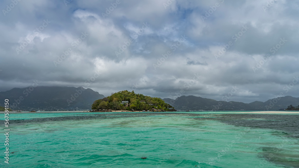 A small island in the turquoise ocean is completely overgrown with tropical vegetation. Villas are visible through the foliage. The boats are moored at the shore. Clouds in the sky. Seychelles
