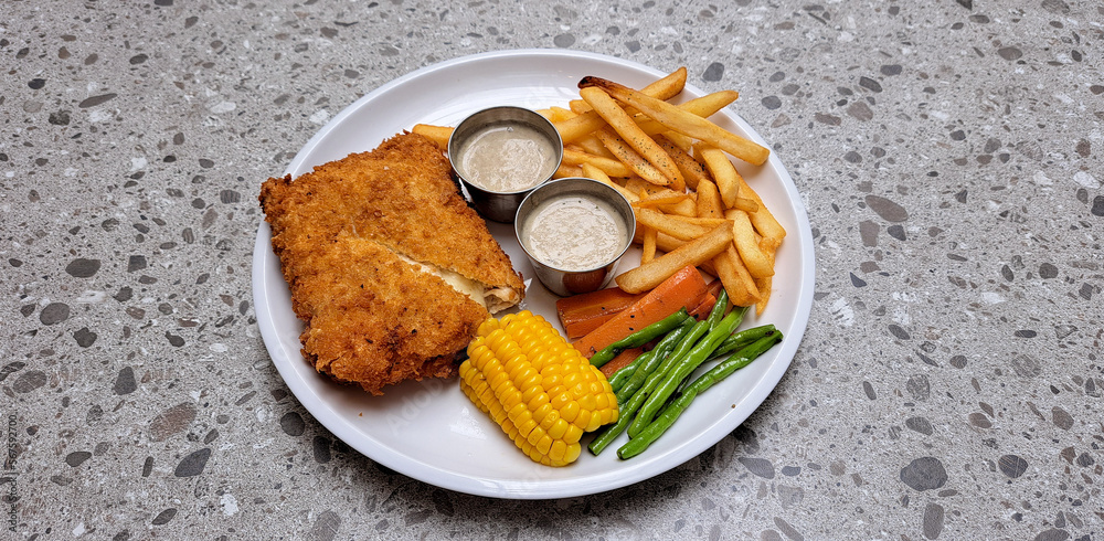 Chicken Cordon Bleu set of plate with potato French fries, corn slash, pepper seasoned carrot & long peas, accompanied with mushroom sauce on a white plate. Served on top of grey terrazzo table.