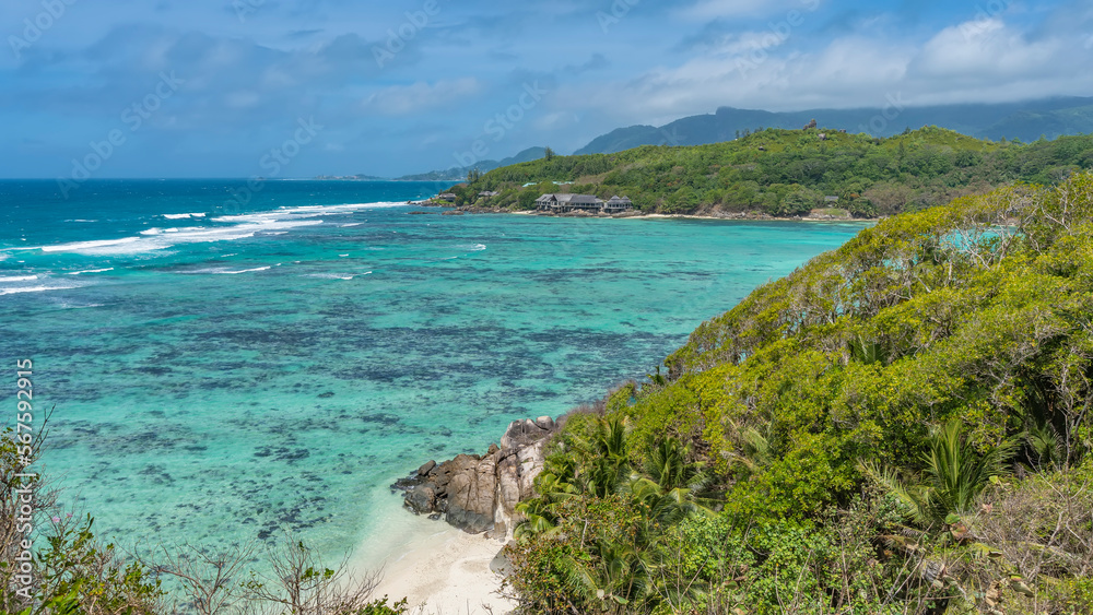 The observation deck offers a view of the turquoise ocean. Corals are visible on the bottom, foam waves on the surface. Lush green vegetation on the hills, boulders near the shore.Blue sky. Seychelles