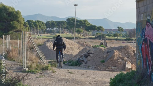 Cyclist cicles trough  buildingsite around sand heaps , wire fence on one side and wall with graffitti on other side, nice landscape with trees and mountains in distance. photo