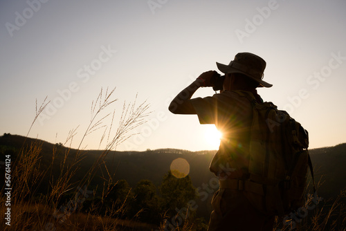 boy scout schoolboy silhouette hiking with backpack standing on a cliff Boy scouts look through binoculars exploring a beautiful forest in the evening as the sun sets. © เลิศลักษณ์ ทิพชัย