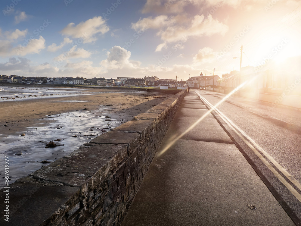 Foot path by Kilkee beach and town houses in the background. County Clare, Ireland. Nobody. Popular summer resort. Atlantic ocean, Irish seascape. Cloudy sunrise sky and sun flare. Calm mood