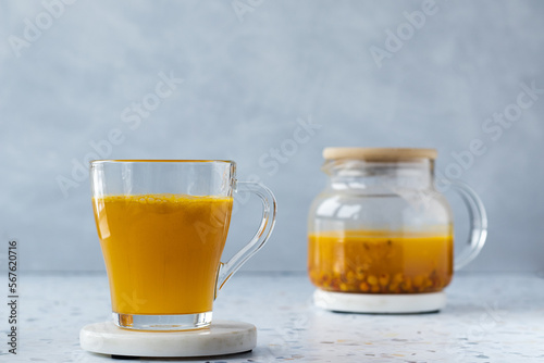 Hot sea ​​buckthorn tea with ginger and honey in a glass cup. Teapot on a terrazzo table. Grey background. Close up. Vitamin drink, immune support, cold remedy. Health supplements. Copy space