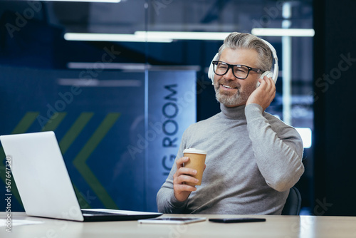 Mature businessman in headphones listening to audio books online podcasts and radio with music, man working inside office at laptop, boss smiling happy with achievement results and work. photo