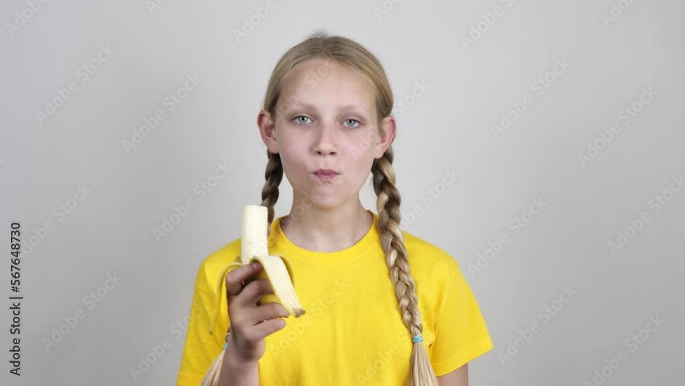 a girl with pigtails bites and chews a ripe banana, looking at the camera in a yellow t-shirt on a white background. Healthy food and beauty a girl with pigtails bites and chews a ripe banana, looking at the camera in a yellow t-shirt on a white background. Healthy food and beauty