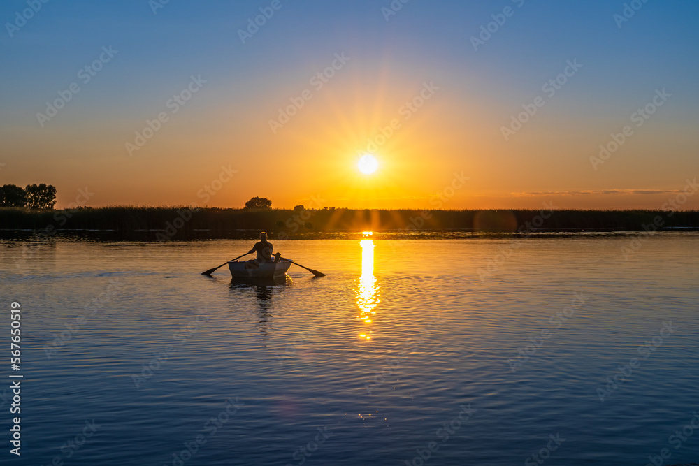 Silhouette of a 60-year-old Caucasian man on a boat with a poodle dog rowing oars to meet the sunset over the lake