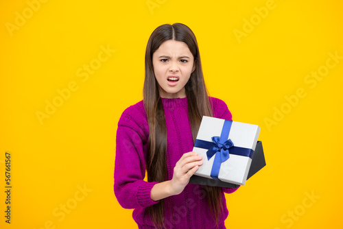 Angry teenager girl, upset and unhappy negative emotion. Teenager child holding gift box on yellow isolated background. Gift for kids birthday. Christmas or New Year present box. © Olena