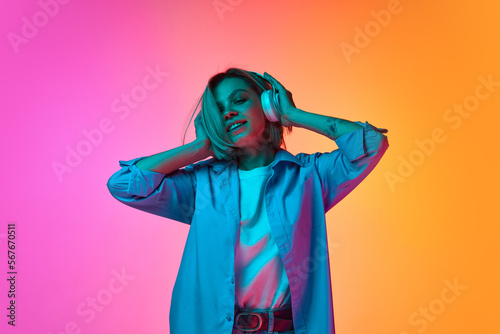 Portrait of young Caucasian happy girl, student in casual style clothes listening to music and dancing over gradient pink-orange background in neon light.