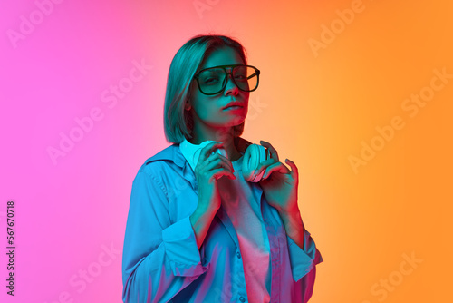 Young stylish girl, student in casual clothes wearing fashionable eyewear posing over gradient pink-orange background in neon light. Fashion, style, ad