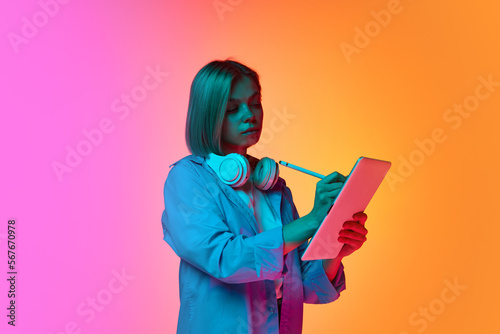 Portrait of young female student using tablet isolated over gradient pink-orange background in neon light. Concept of education, online studying, active lifestyle, youth, emotions