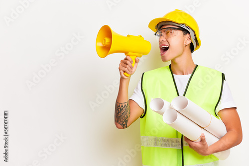 Horizontal shot of distressed Asian handyman holds megaphone screams loudly has angry expression poses with blueprint wears protective glasses helmet and uniform isolated over white wall. People photo