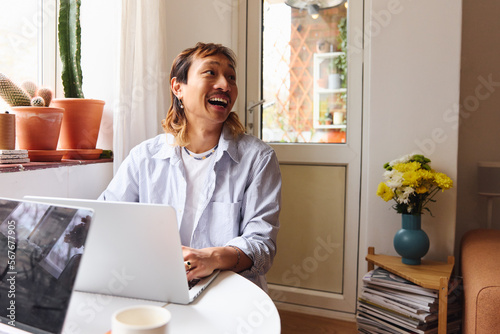 Close Up Of Asian Man In Lounge Sitting At Table With Laptop Working From Home Laughing off Camera photo