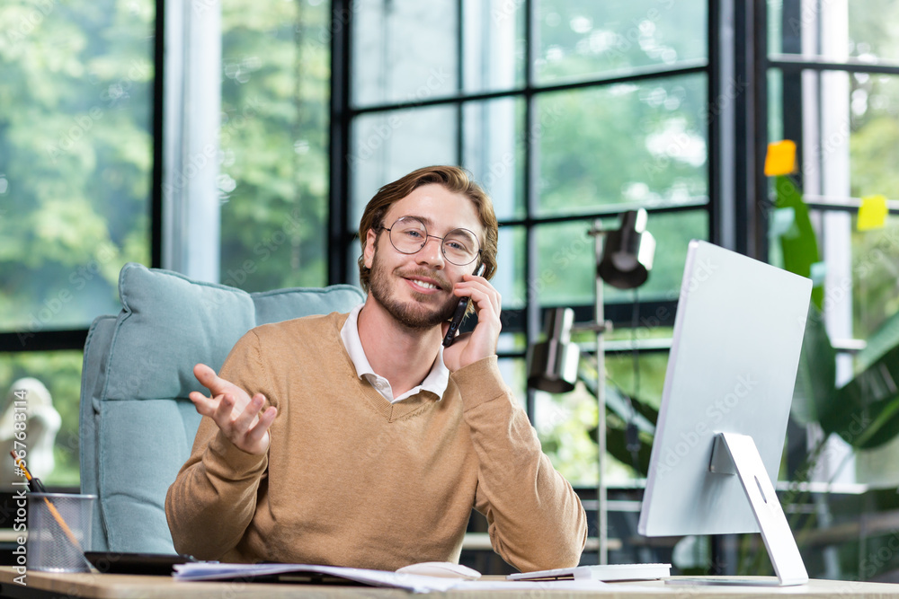 Young handsome man businessman, freelancer sitting in the office at the computer and talking on the phone. Looks into the camera. gesturing, smiling. Young handsome man businessman, freelancer sitting in the office at the computer and talking on the phone. Looks into the camera. gesturing, smiling.