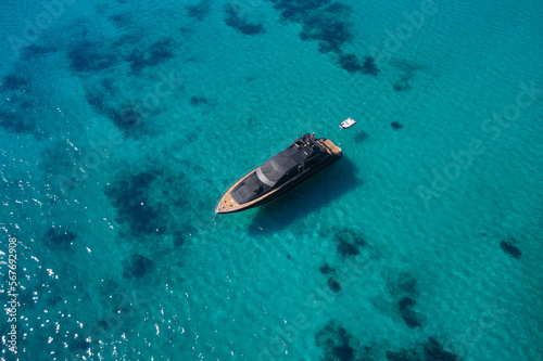 Dark blue yacht with teak wood in the morning sun top view. Large innovative modern yacht anchored on clear water aerial view.