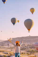 Happy traveller girl raising hands while watching magnificent view of flying hot air balloons in famous tourist attraction - Cappadocia