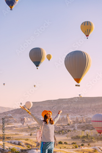 Happy traveller girl raising hands while watching magnificent view of flying hot air balloons in famous tourist attraction - Cappadocia