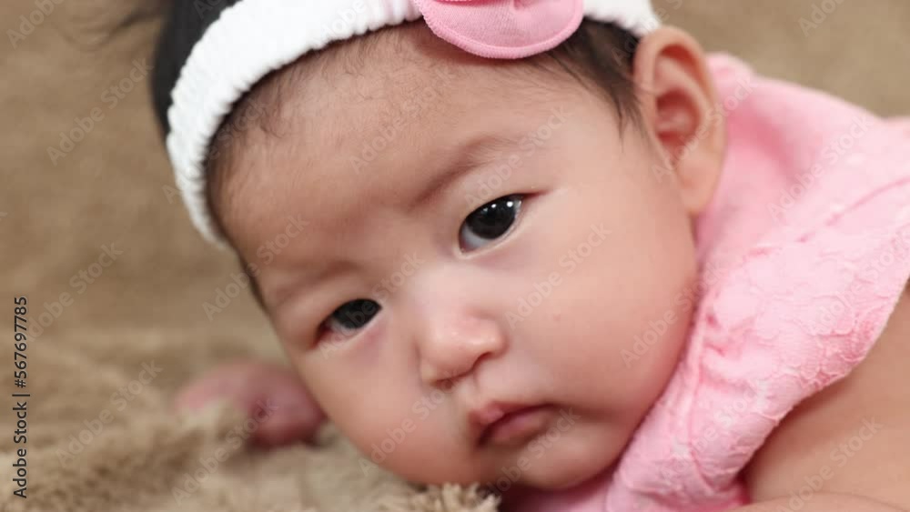 Closeup Asian newborn curiosity in beautiful pink dress prone down on sofa, infant girl looking for mom, healthy toddler want to play with mother while she work in bedroom, baby feel hungry want milk. Closeup Asian newborn curiosity in beautiful pink dress prone down on sofa, infant girl looking for mom, healthy toddler want to play with mother while she work in bedroom, baby feel hungry want milk.