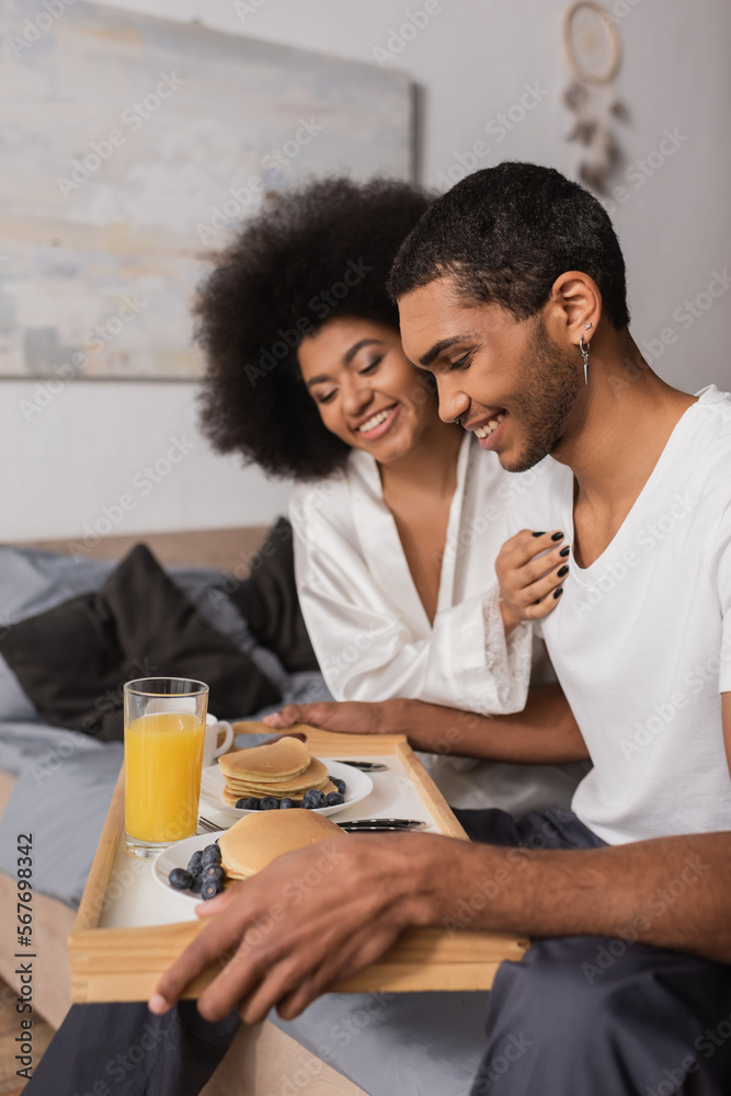 happy african american man holding tray with pancakes and drinks near sexy girlfriend in bedroom happy african american man holding tray with pancakes and drinks near sexy girlfriend in bedroom