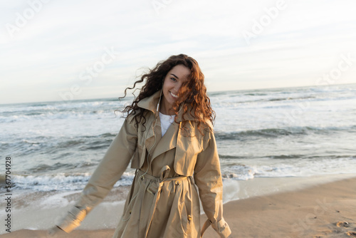 cheerful young woman in beige trench coat and wired earphones listening music near sea in Barcelona.