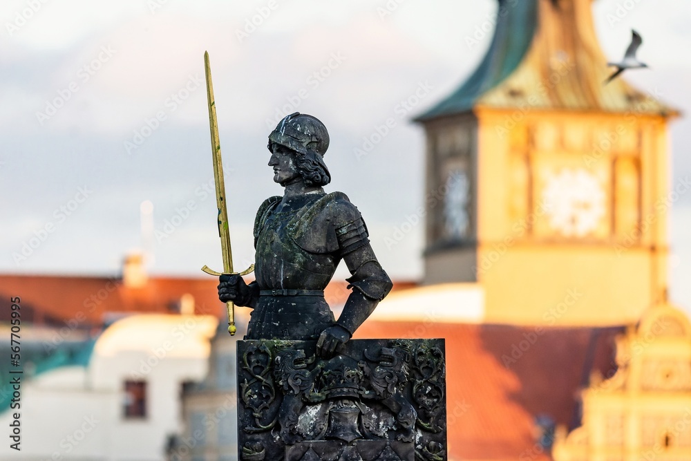Prague, Czech Republic - December 8 2022: View of Brunswick statue, a Czech knight, holding a golden sword and coat of arms. Sunny evening, blue sky and a yellow water tower in the background.
