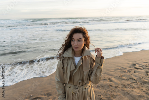 Young woman in trench coat touching curly hair on beach.