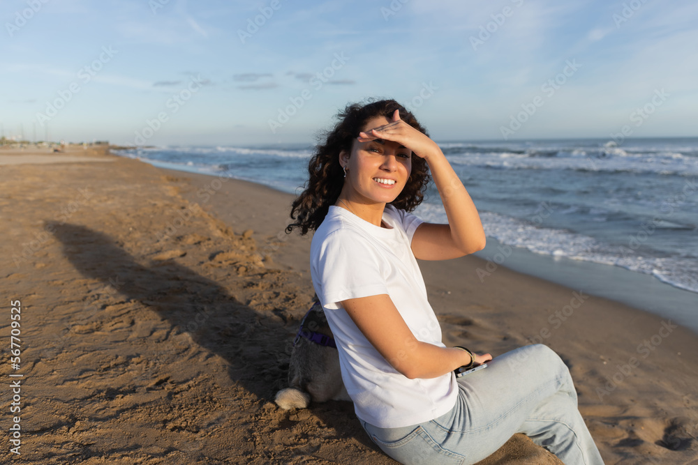 cheerful woman in white t-shirt holding smartphone and sitting with pug dog on beach in Barcelona. cheerful woman in white t-shirt holding smartphone and sitting with pug dog on beach in Barcelona.