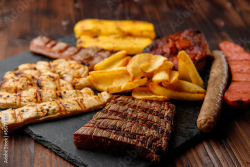 top view of typical Italian tyrolese appetizer of grilled meat mix, homemade potato chips, polenta on black stone dish on wooden background