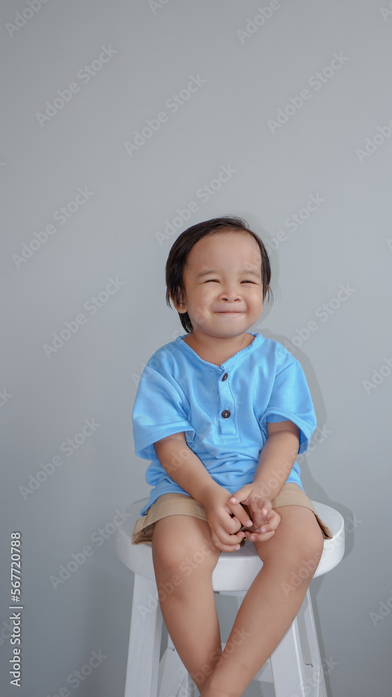 cute little asian boy sitting at stool and smiling to the camera, on grey background. cute little asian boy sitting at stool and smiling to the camera, on grey background.