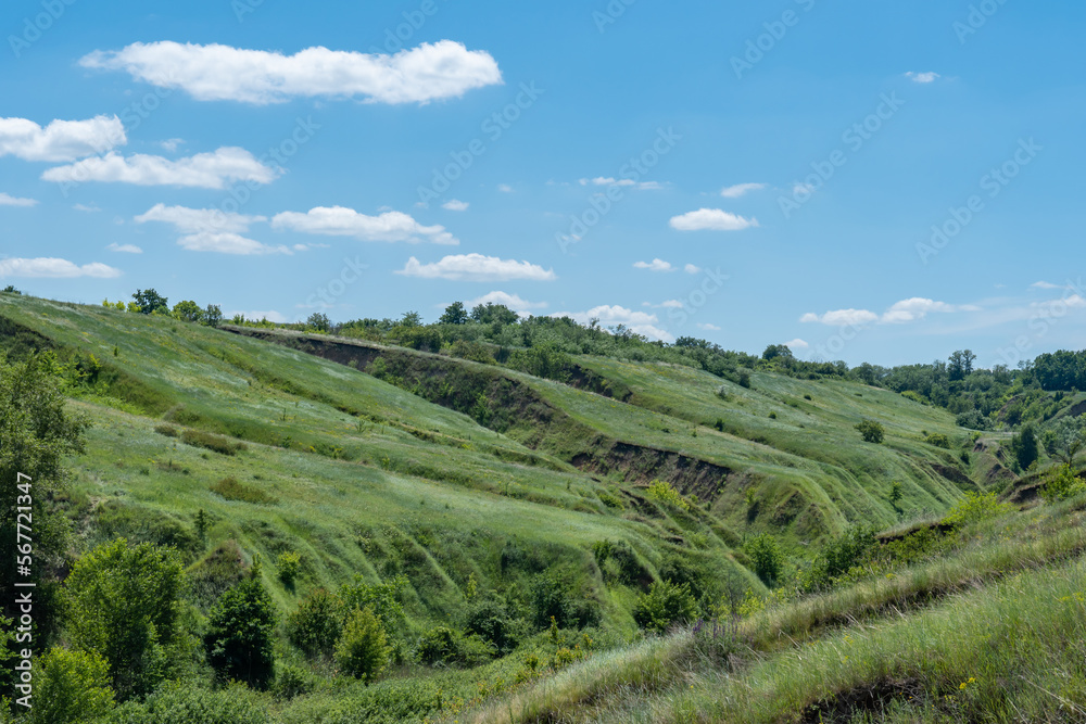 View ravine covered with greenery. Landscape valley with geological faults. Earthen mountains and bumps relief on against sky. Hill beam with protrusions and recesses. Grass and trees on slope canyon.