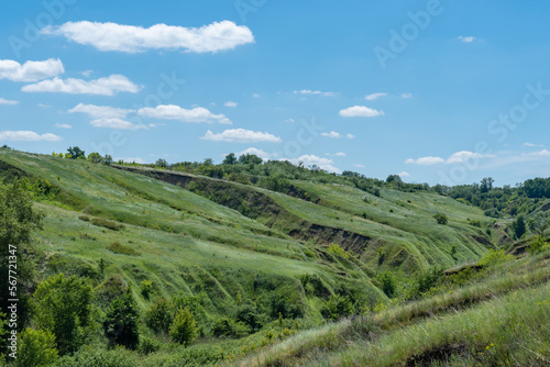 View ravine covered with greenery. Landscape valley with geological faults. Earthen mountains and bumps relief on against sky. Hill beam with protrusions and recesses. Grass and trees on slope canyon.